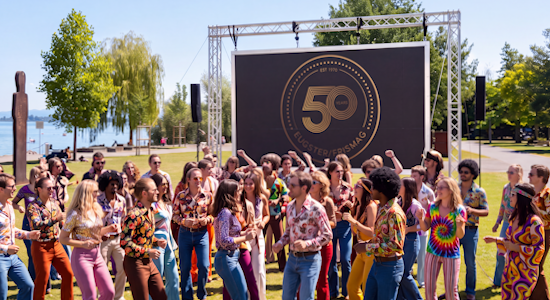 LED-Screen Miete für Firmenjubiläum und Outdoor-Events (2) Großer mobiler LED-Screen auf einer Wiese am See bei einem Firmenjubiläum im 70er-Jahre-Stil, auf dem das Logo „50 Years Eugster/Frismag“ angezeigt wird, während Menschen in Retro-Kleidung davor tanzen.