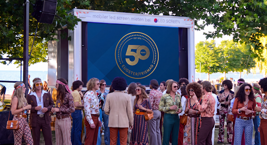 Mobiler LED-Screen Trailer für Outdoor-Events mieten Mobiler LED-Screen Trailer von LED-SCREEN an einer Strandpromenade bei einem Firmenjubiläum im 70er-Jahre-Stil, auf dem Bildschirm läuft das Logo „50 Years Eugster/Frismag“.